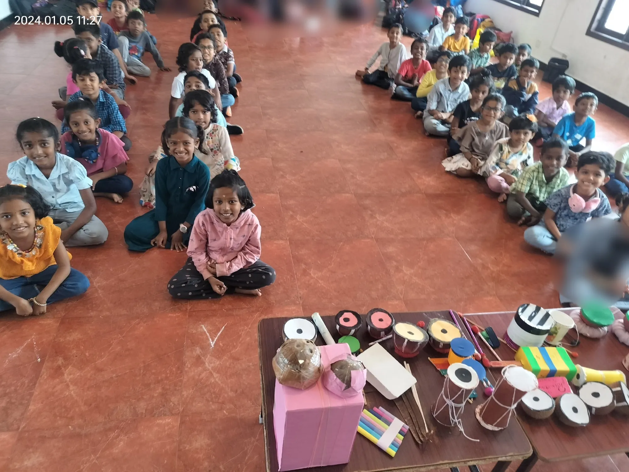 A joyful assembly of 1st graders sitting cross-legged, showcasing their eco-friendly musical instruments at Vidhya Peetham i-School during the harmonious echoes activity. ??