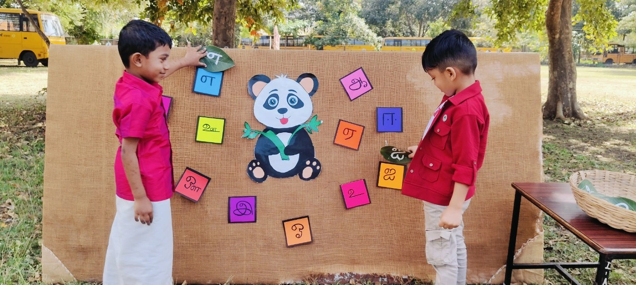 Students in red attire practicing Tamil letter recognition with panda display