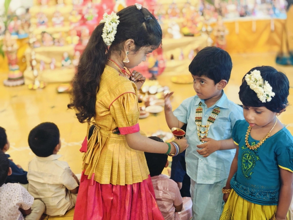 A girl in a silk pavadai offering kumkum to a small boy in front of the Navaratri golu