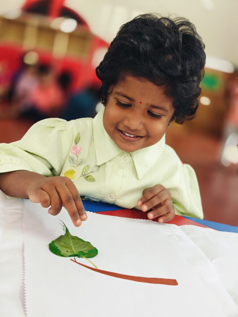A smiling child places a green leaf face down on white paper at the end of a brown painted line.