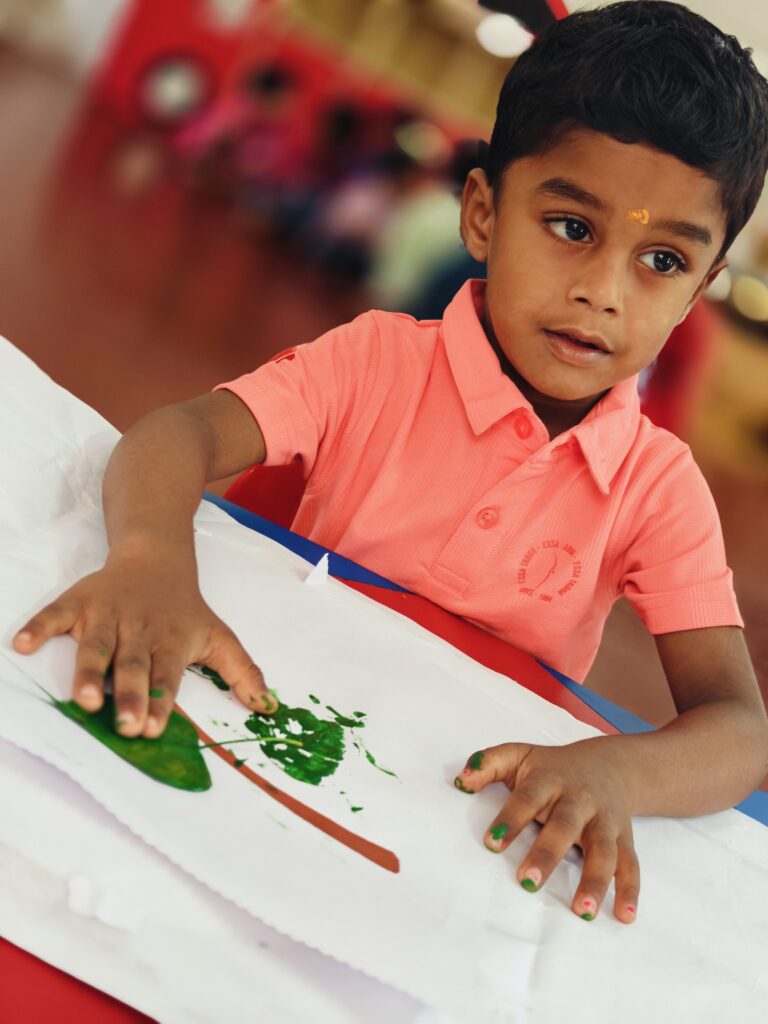 A child presses a painted leaf onto white paper, with green paint on the fingertips and a print beside a brown line.