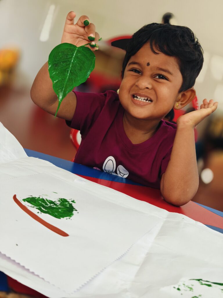 A grinning child with green-painted fingertips holds up a paint-covered leaf above a fresh leaf print.