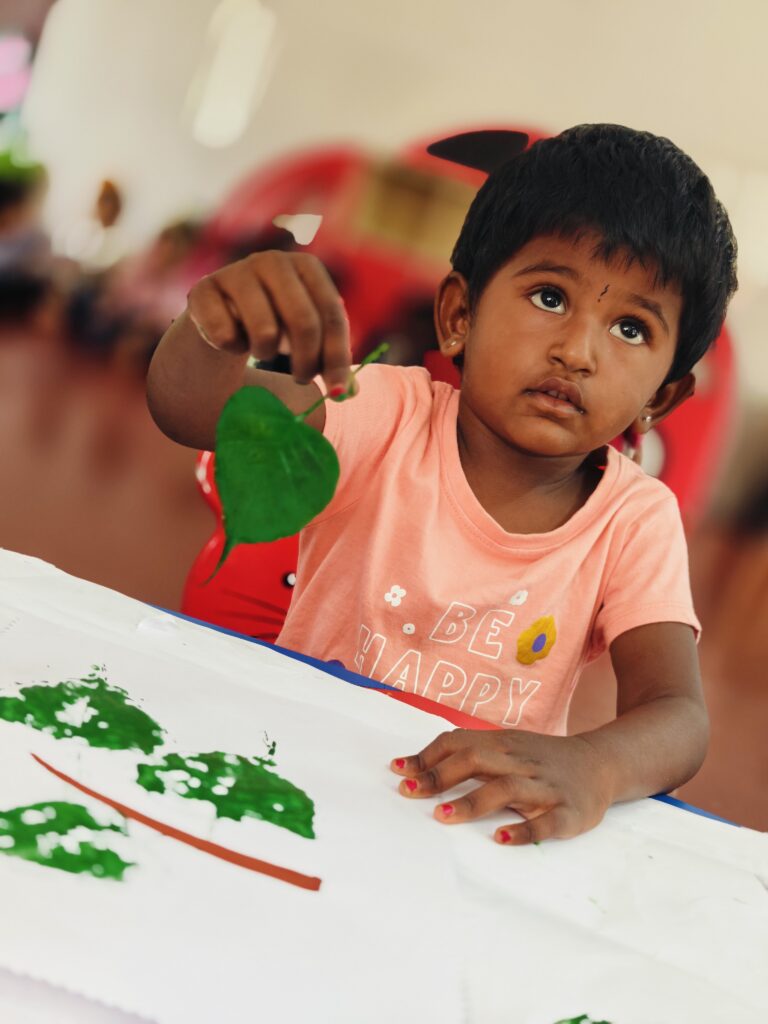 A child holds up a green leaf above a sheet of paper that already has three green leaf prints on it.