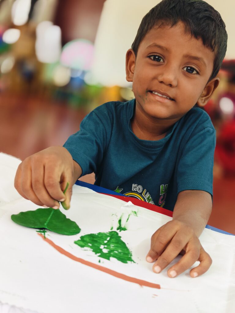 A child smiles at the camera while pressing a green-painted leaf onto paper next to an earlier leaf print.