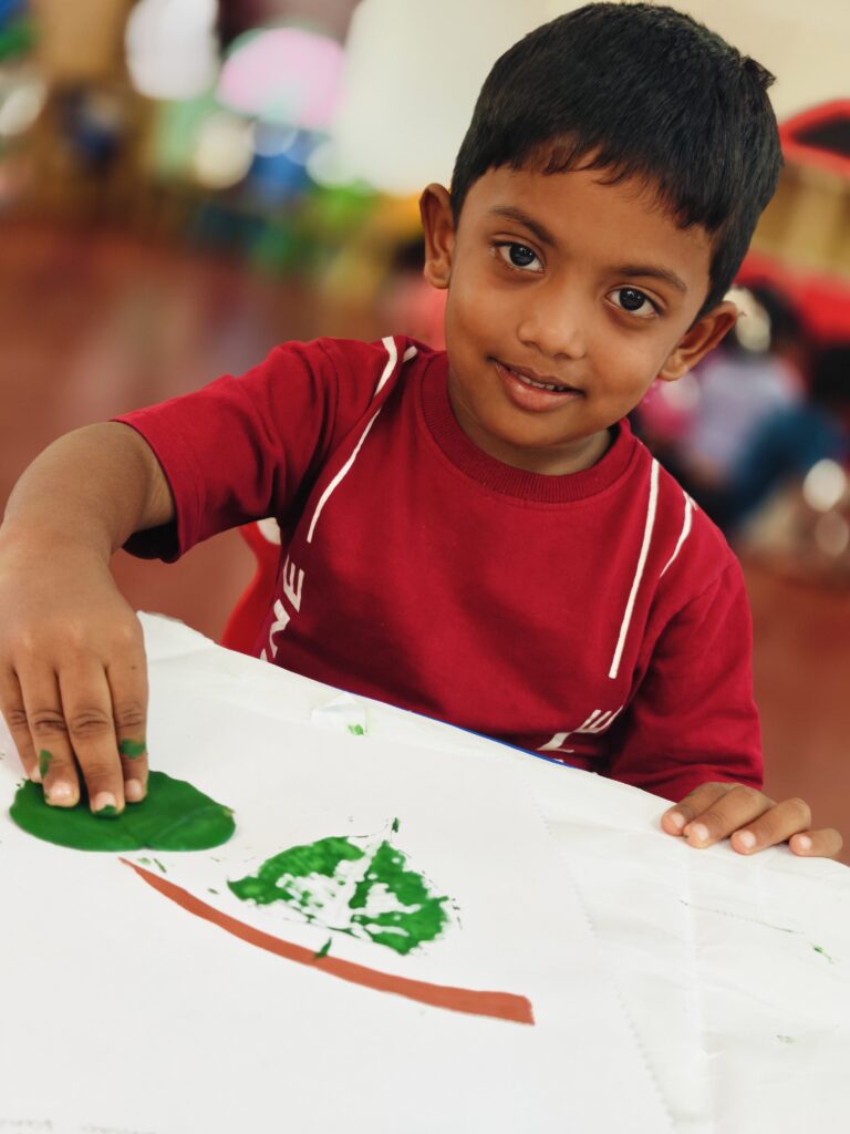 A child in a red T-shirt presses a painted leaf onto paper beside a green leaf print and a brown line.