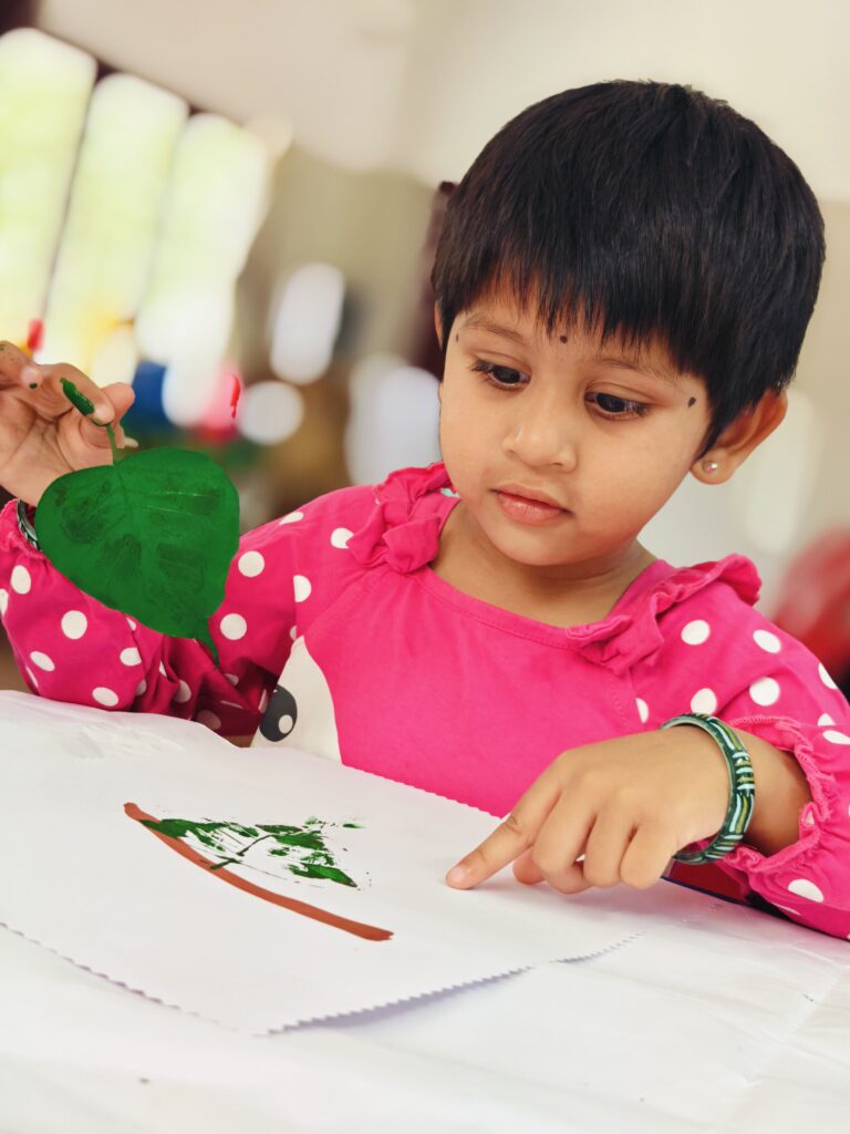 A child in a pink polka-dot top holds a painted leaf and points to a green leaf print on white paper.