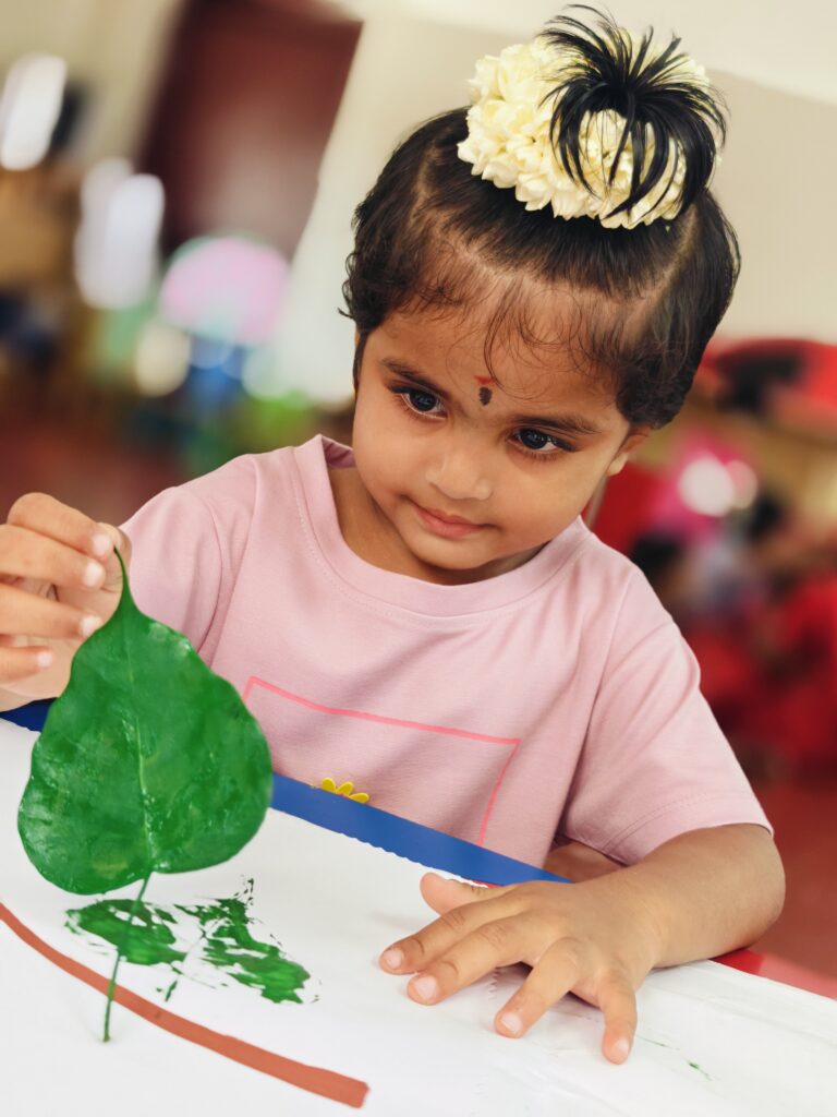 A child in a pink T-shirt lifts a painted leaf from paper, leaving a green print beside a brown painted line.