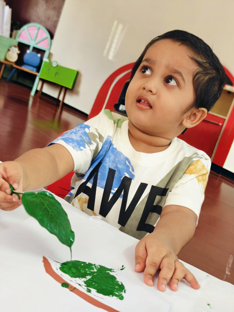 A child looks up while lifting a painted leaf from paper, showing a green print in a bright classroom.