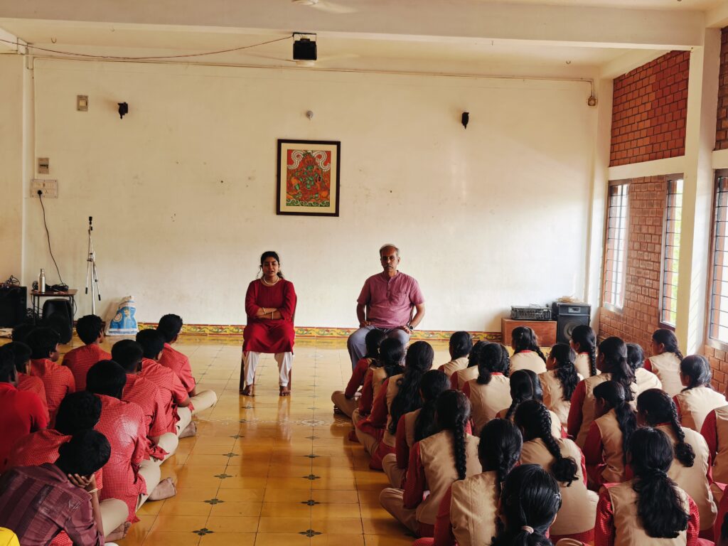 Wide view of the hall with two speakers seated beneath a framed painting and students seated on the floor.
