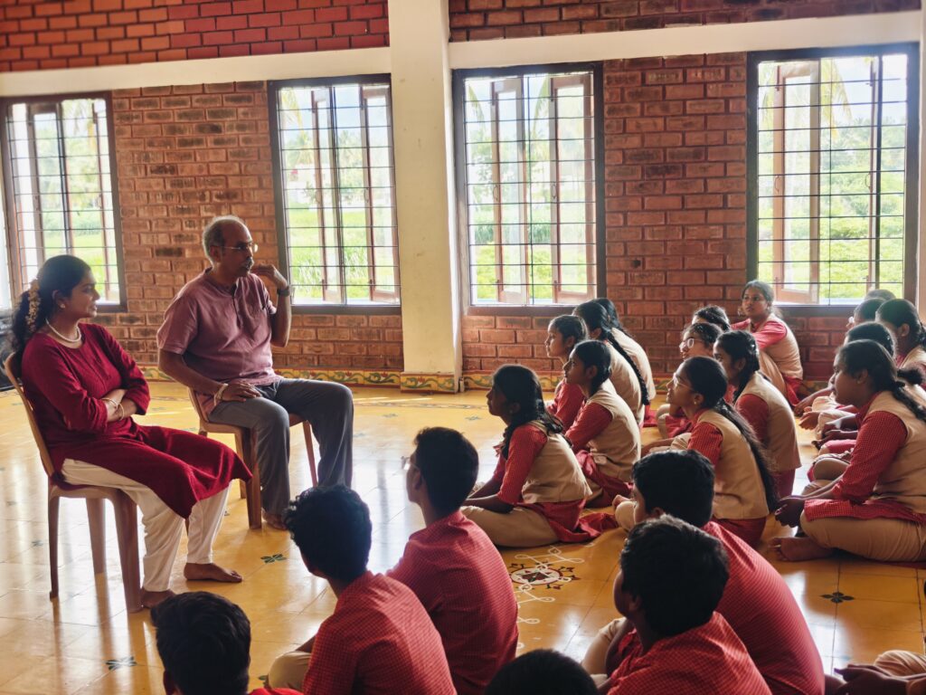 A woman in a maroon kurta and a man in a mauve shirt sit on chairs, speaking to students seated on the floor.