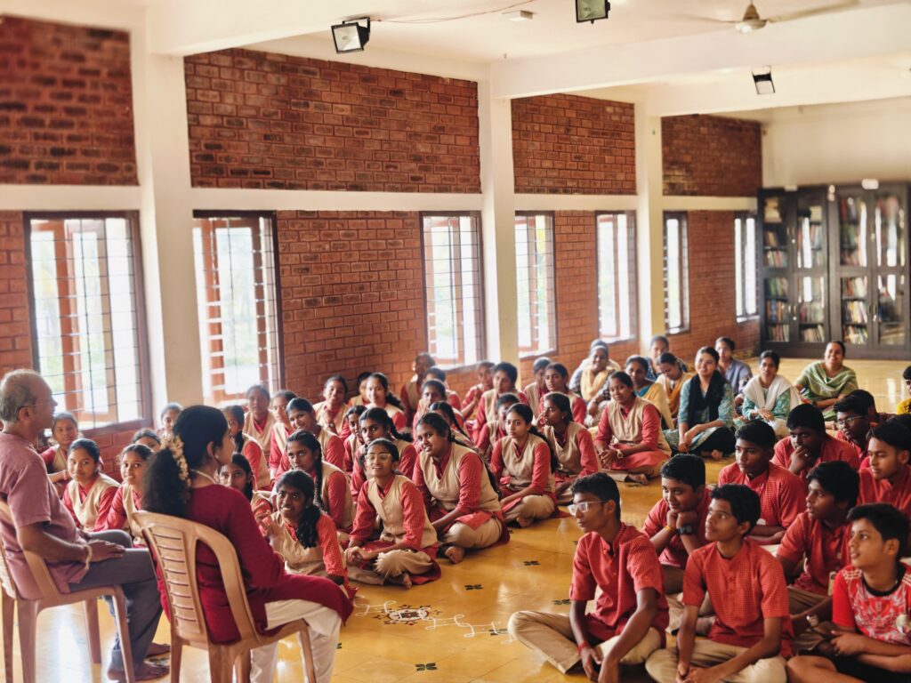 A large group of students sits on the floor of a brick-walled hall, listening to two seated speakers.