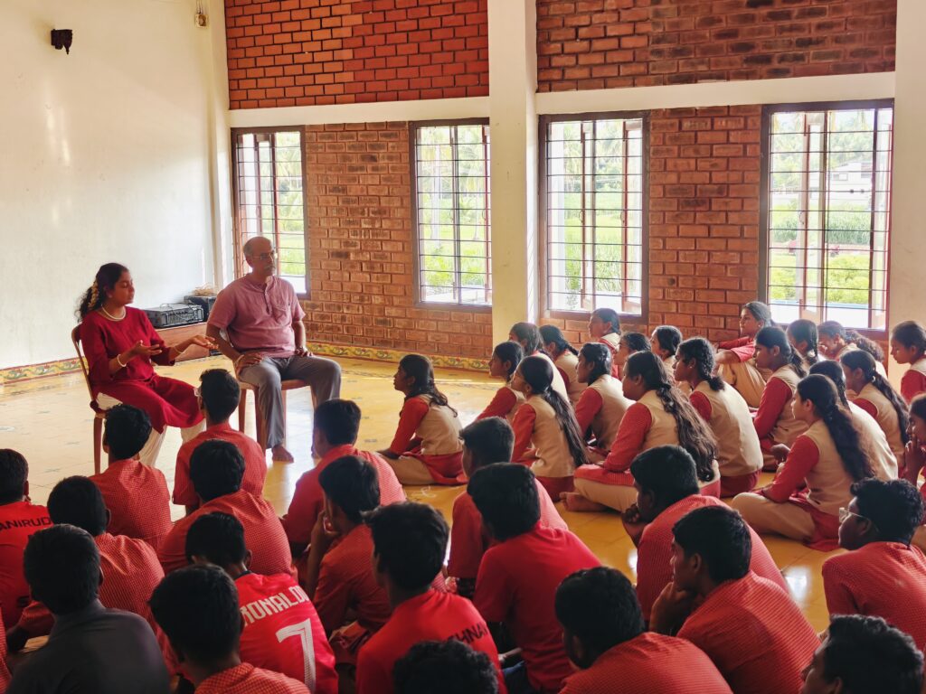 Students sit on the hall floor facing two seated speakers in front of a white wall and brick windows.
