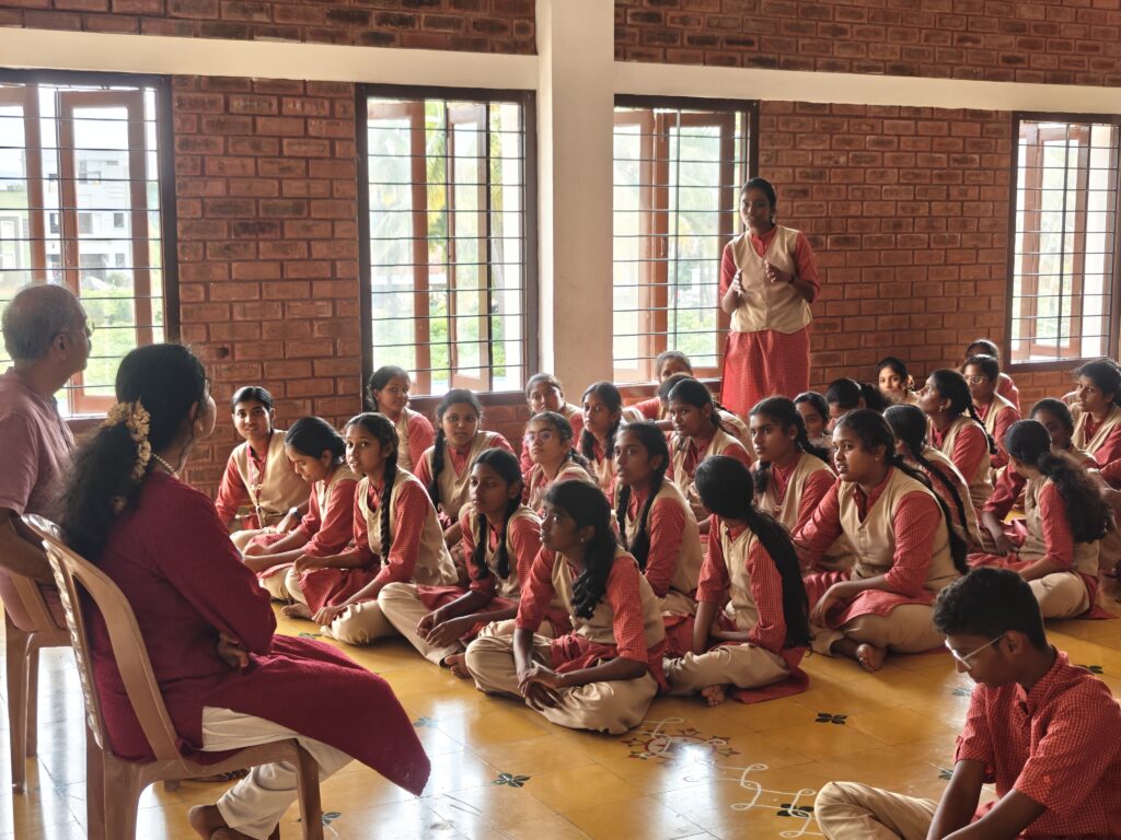 A student stands among seated classmates to speak, while two adults on chairs listen.