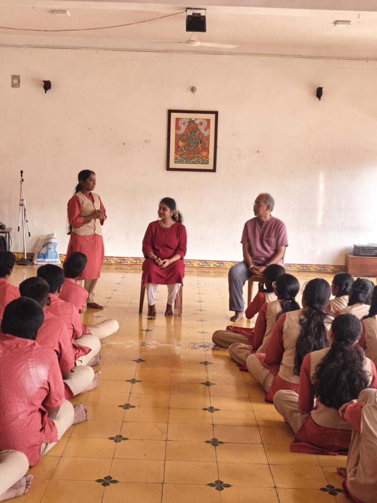 A student in uniform stands to speak while two adults sit on chairs beside her and students listen.