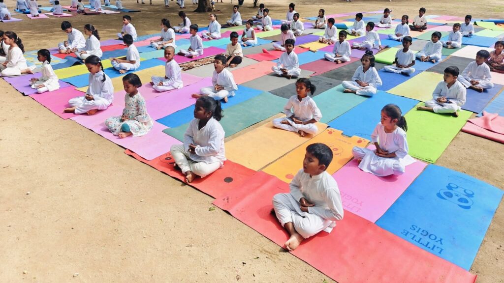 Children sit cross-legged with hands on knees on rows of colourful mats outdoors.