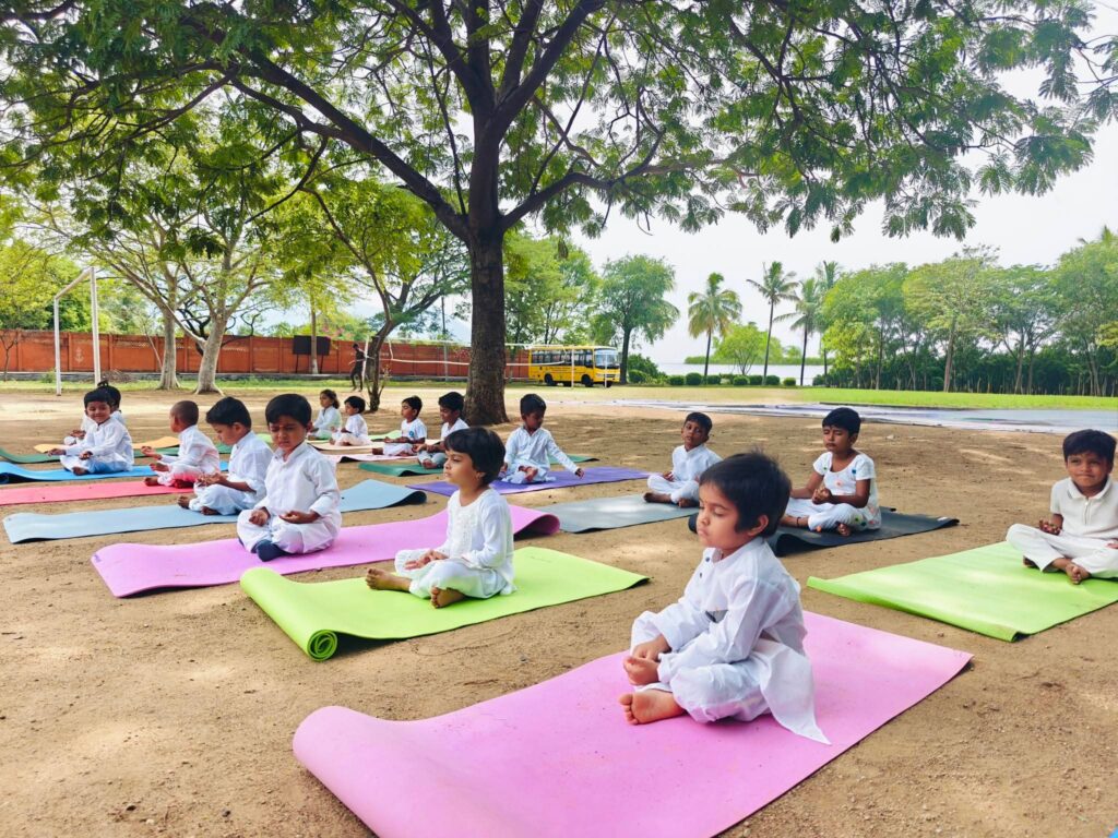 Very young children sit cross-legged with eyes closed on mats under a large tree.