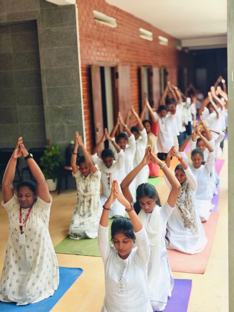 Students and an adult kneel with arms raised and palms joined above their heads in a corridor.