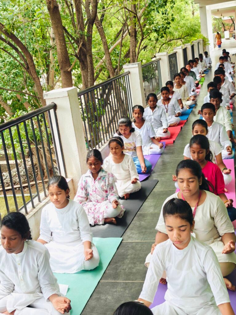 Students sit cross-legged with eyes closed on mats along a shaded veranda beside trees.