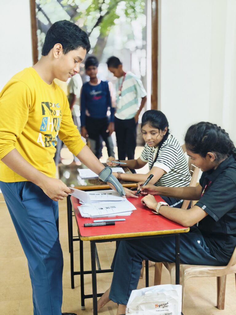 A student stands at the polling desk with ballot papers while a seated student holds a marker pen near his hand.