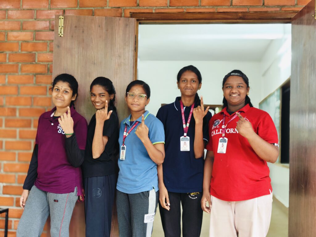 Five girls stand in front of a brick wall and open doorway, each holding up an index finger.