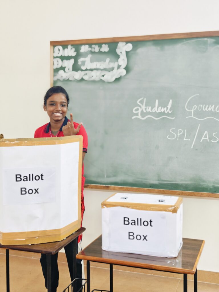 A smiling student holds up her index finger beside two labelled ballot boxes in front of a chalkboard.