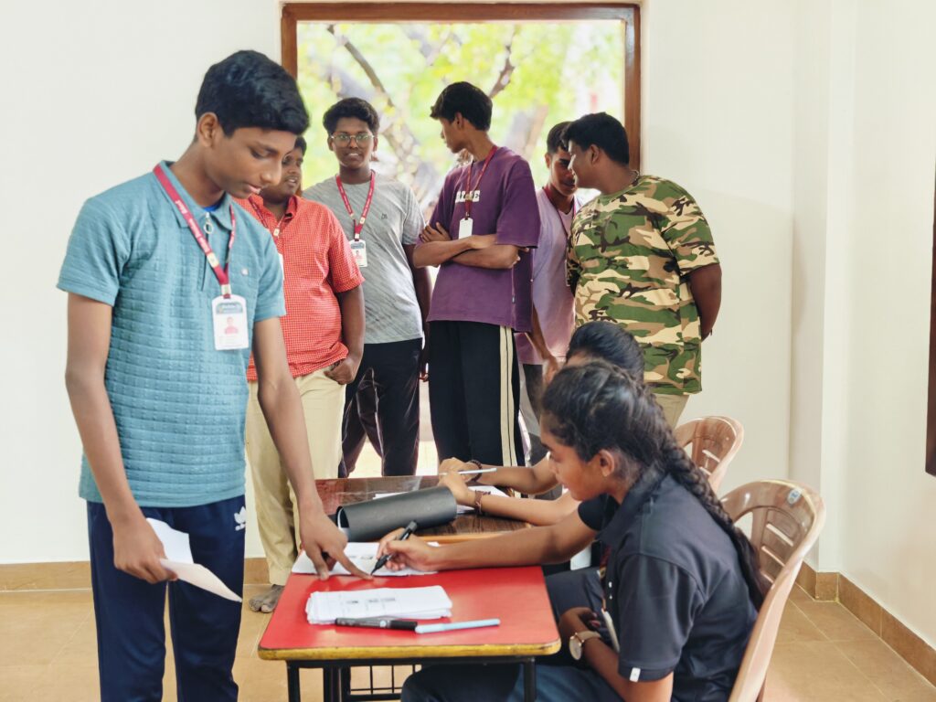A student points to a sheet at a polling desk where two seated students handle ballot papers as others wait behind.