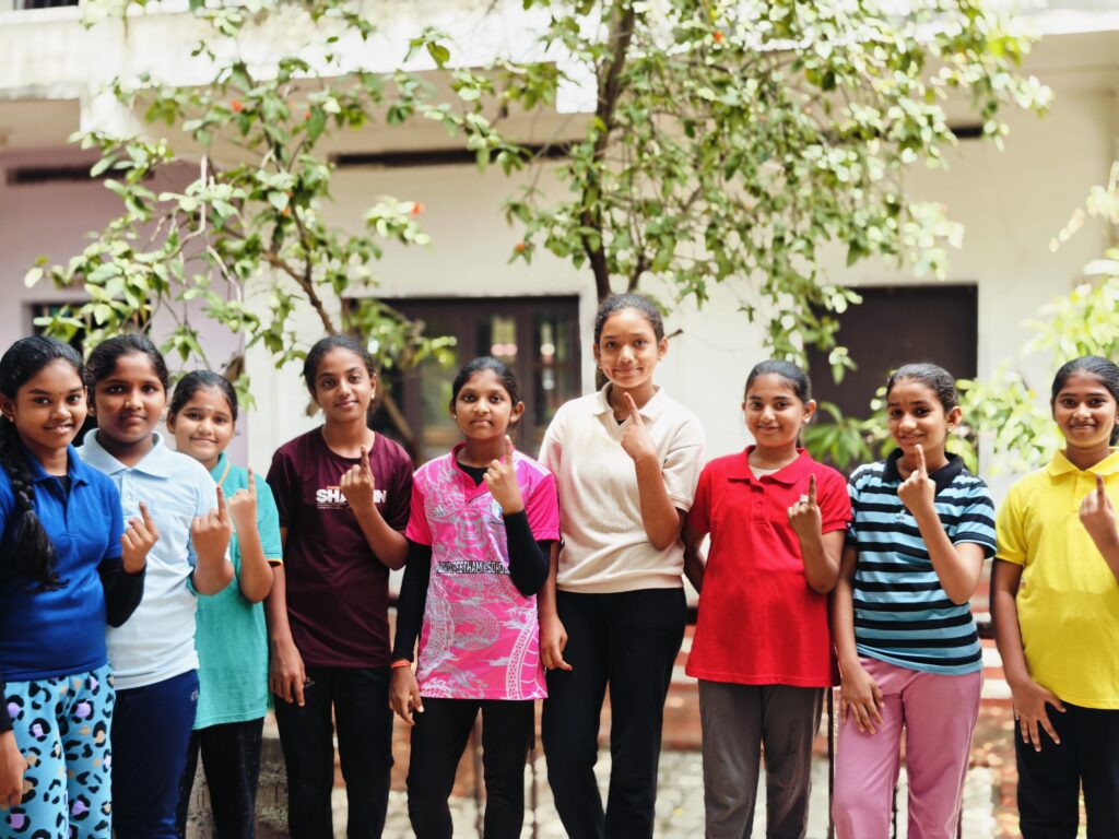 A group of girls stands in a courtyard beneath a tree, each holding up an index finger.