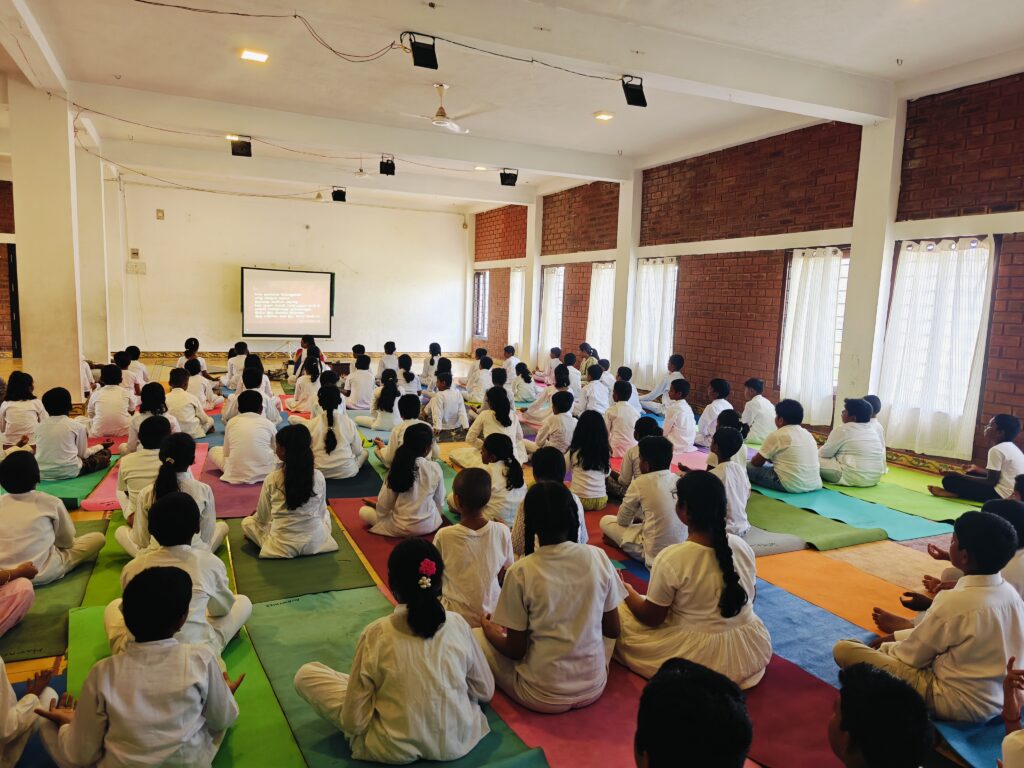Students sit cross-legged on mats in an indoor hall with brick walls, facing a projector screen.