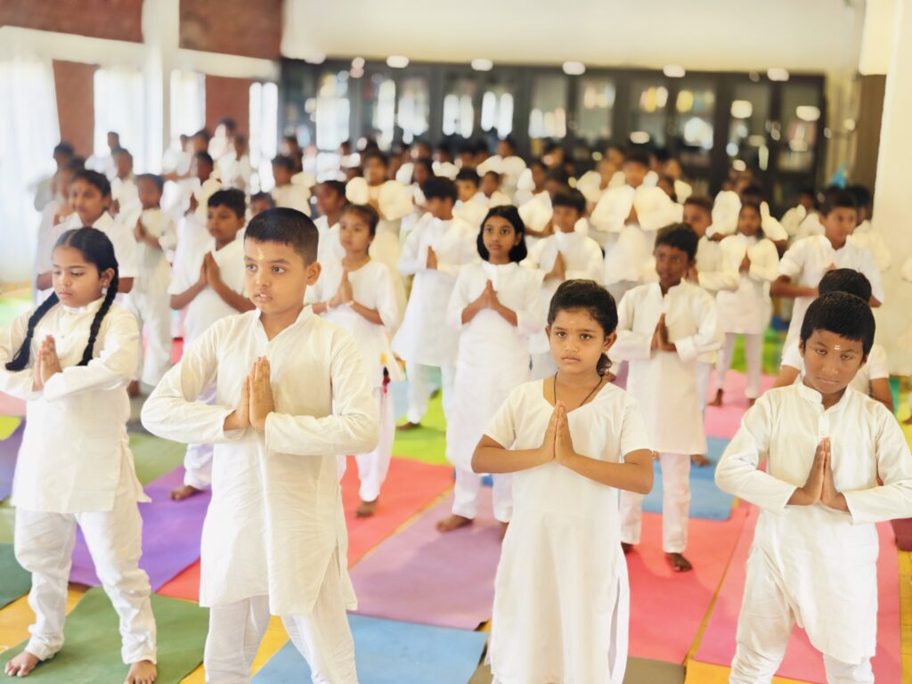 Students in white stand with palms pressed together on colourful mats in an indoor hall.