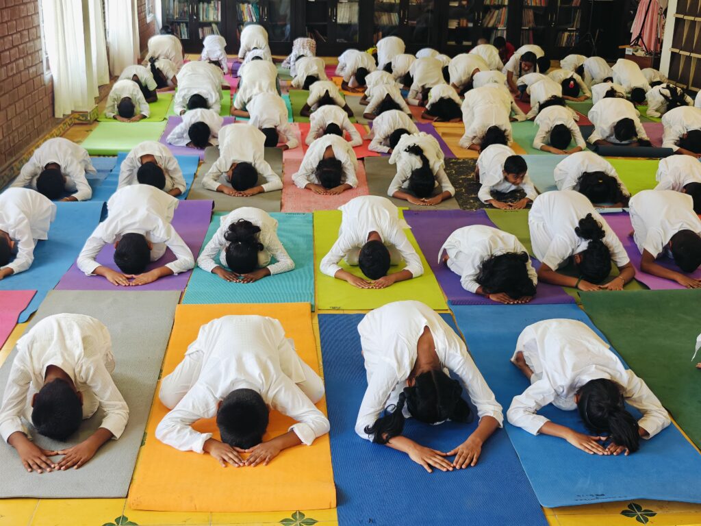 Rows of students kneel and fold forward with foreheads down and hands in front on mats in a hall.