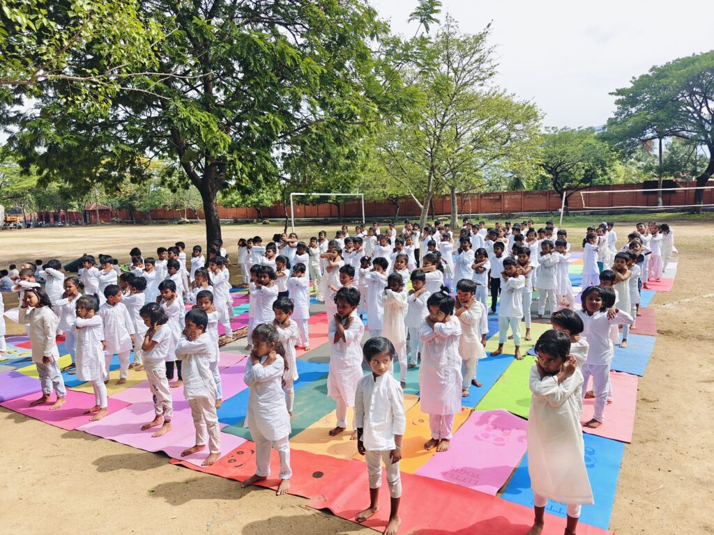 Young children in white stand on colourful mats on the school ground for International Day of Yoga, arms folded.