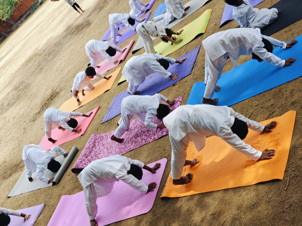 Children in white bend forward with hands and feet on mats outdoors, hips raised in an inverted V shape.
