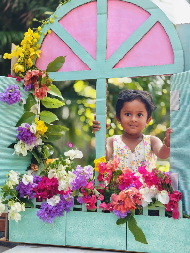 A child in a floral dress stands with both hands on a cardboard window frame lined with bright flowers.