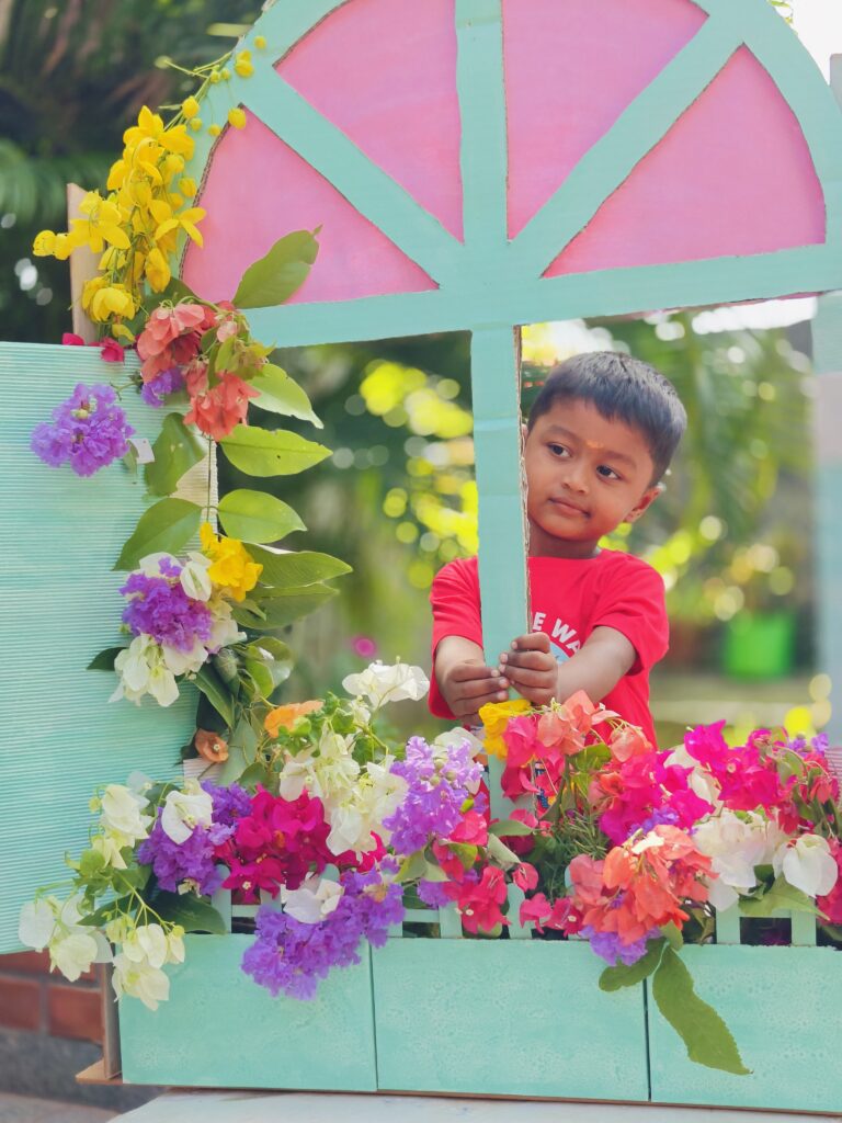 A child in a red T-shirt holds the window frame, surrounded by bougainvillea and yellow flowers.