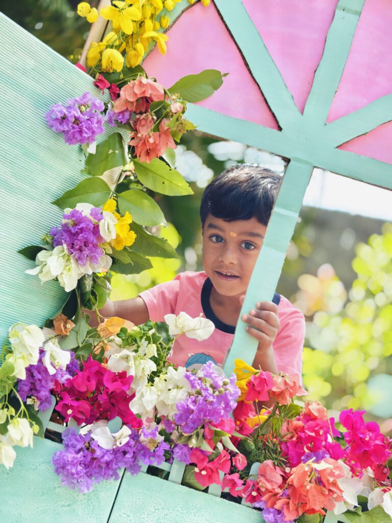 A smiling child in a pink T-shirt peeks around the mint frame of a flower-decorated window prop.