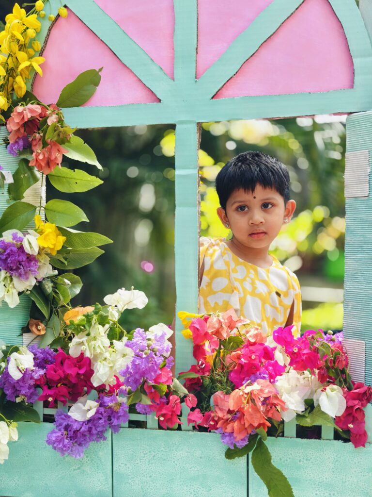 A child in a yellow and white dress looks out from a cardboard window framed with bright flowers.