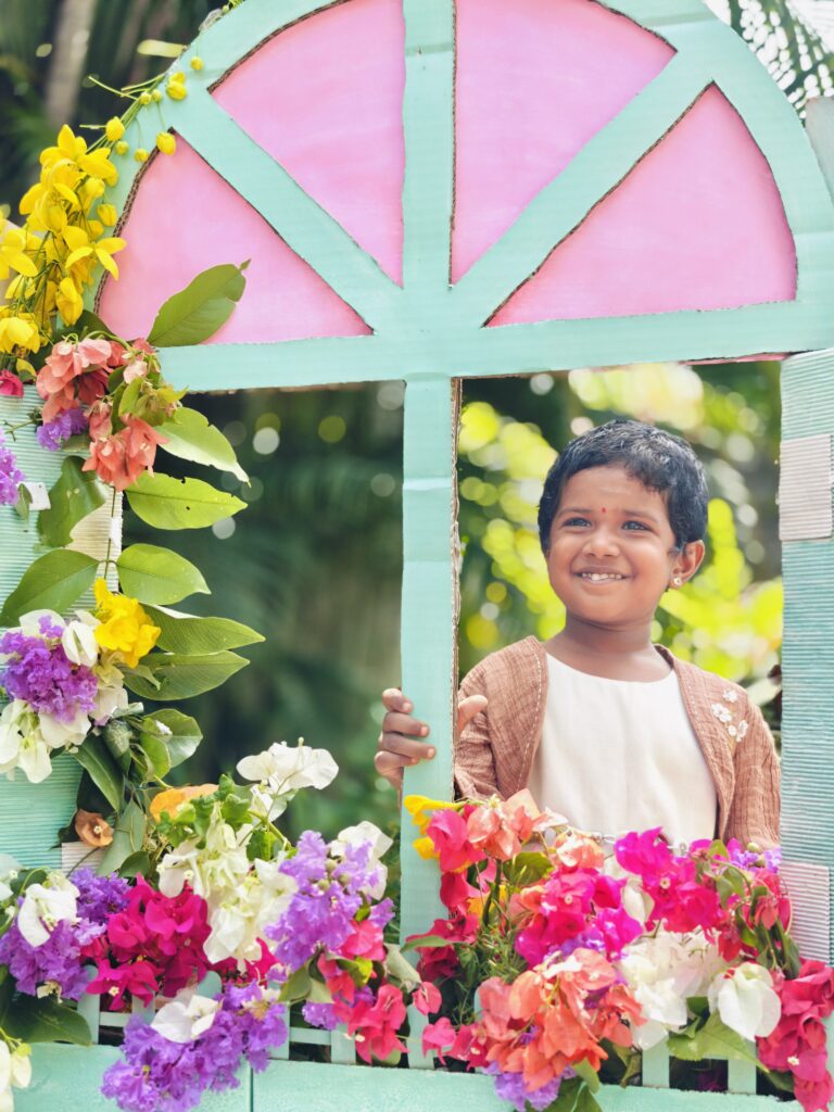 A smiling child in a brown cardigan holds the frame of a pink and mint cardboard window decorated with flowers.