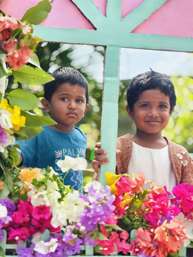 Two children stand together behind a flower-lined cardboard window, one of them smiling.