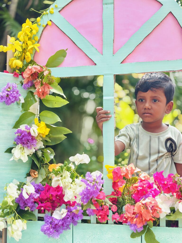 A child in a striped T-shirt holds the window frame, with colourful bougainvillea in front.