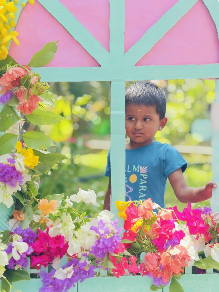 A child in a blue T-shirt looks to the side through a flower-filled cardboard window prop.