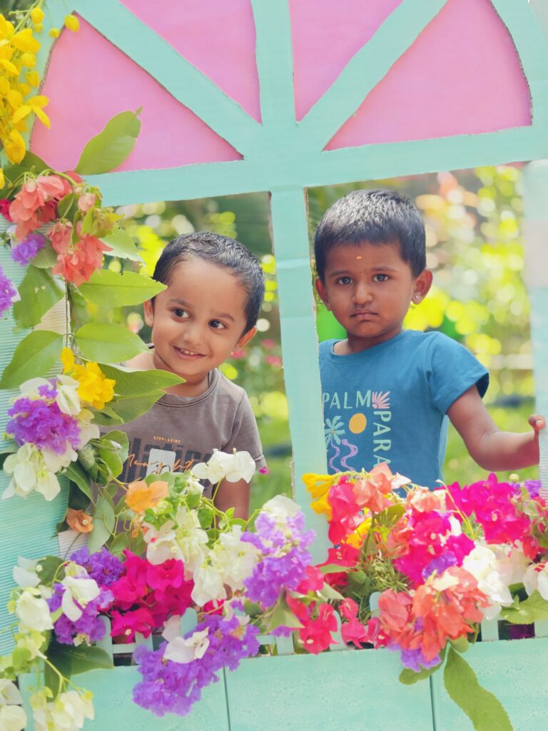Two children, one smiling, look out through a pink and mint cardboard window filled with flowers.