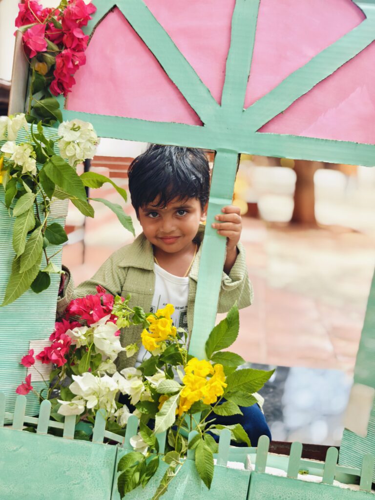 A child in a green jacket smiles behind a window prop decorated with pink, white and yellow flowers.