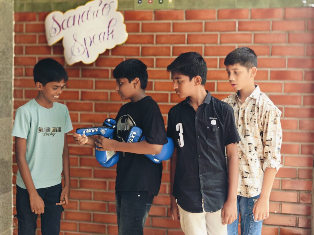 Four boys stand together as one holds a pair of blue boxing gloves during their scenario.