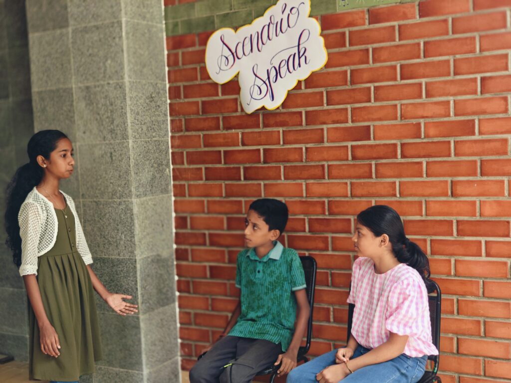 A standing student gestures while speaking to two seated classmates under the Scenario Speak sign.
