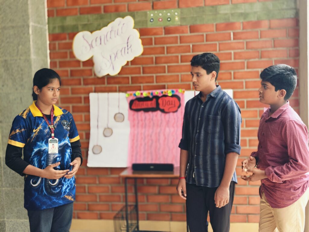 Three students talk in front of a display board reading Annual Day, with a speaker on the table.