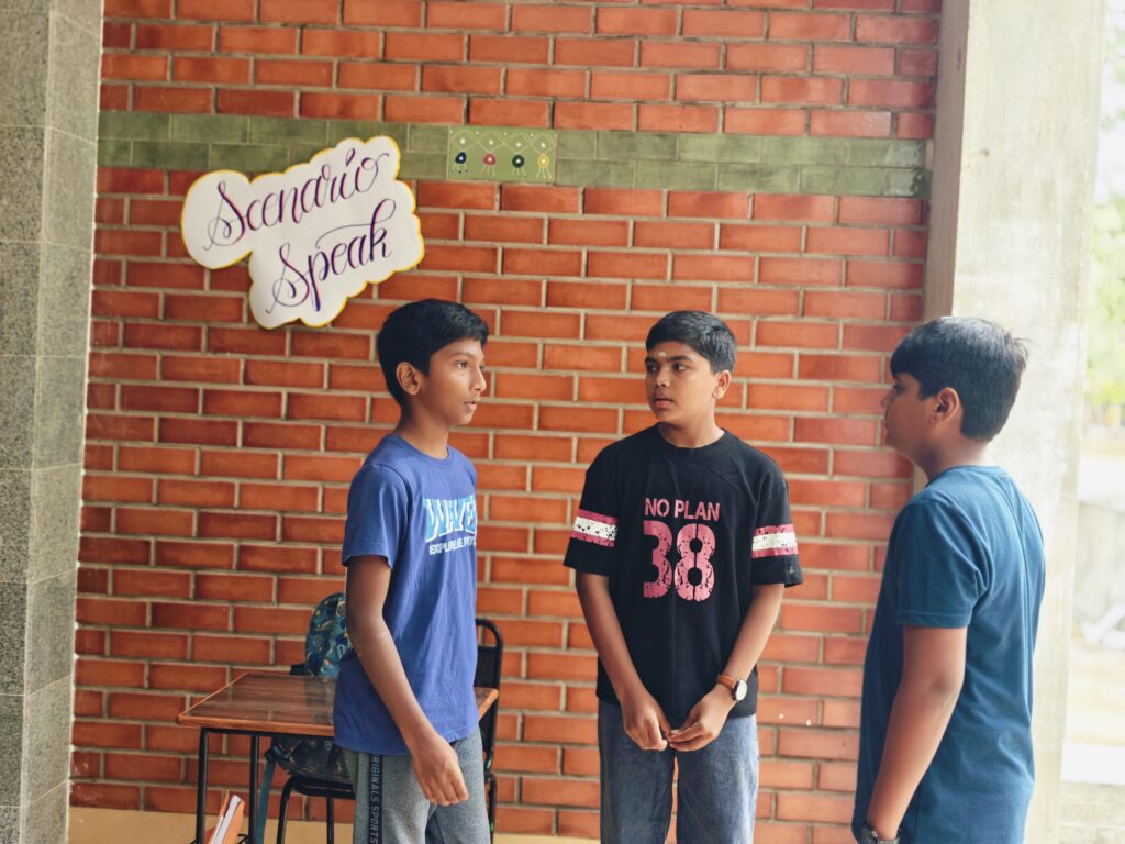 Three boys talk together in front of a brick wall, with a small table and chair behind them.