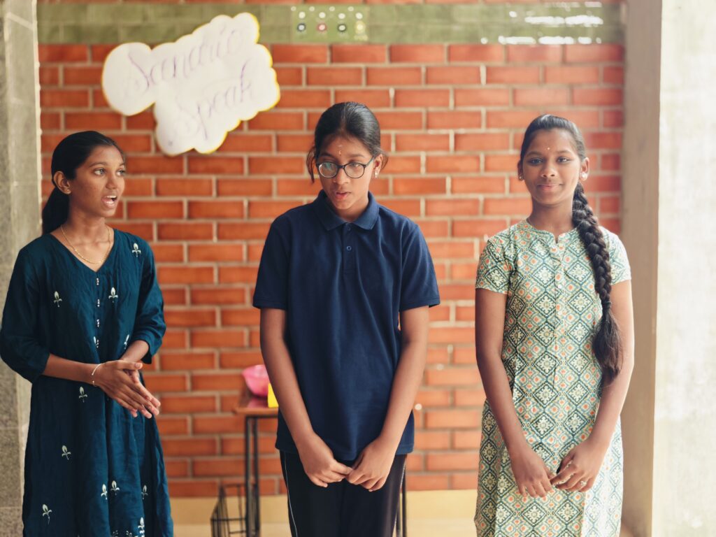 Three girls stand side by side speaking, with a table of props behind them against the brick wall.