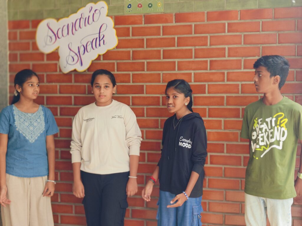 Four students stand in a row against a brick wall, two of them speaking during their scenario.