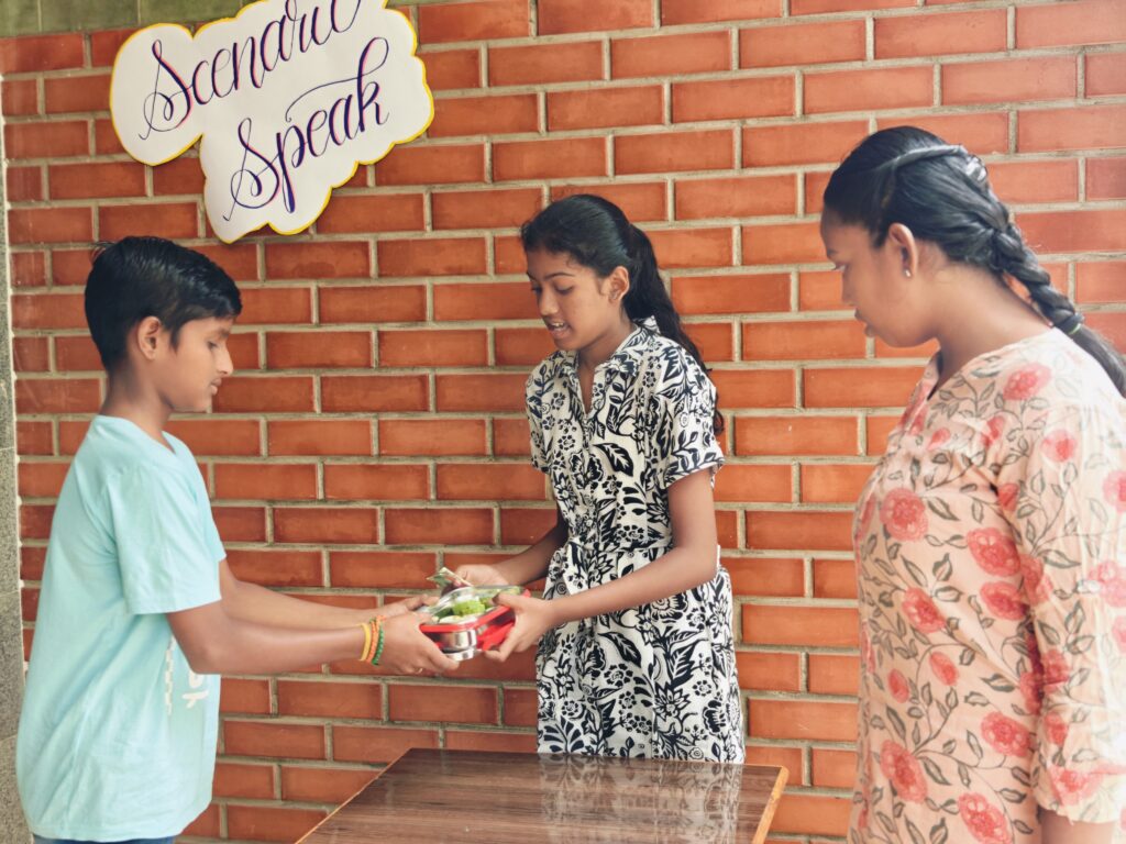 A student hands a lunch box to a classmate across a table while another student watches.