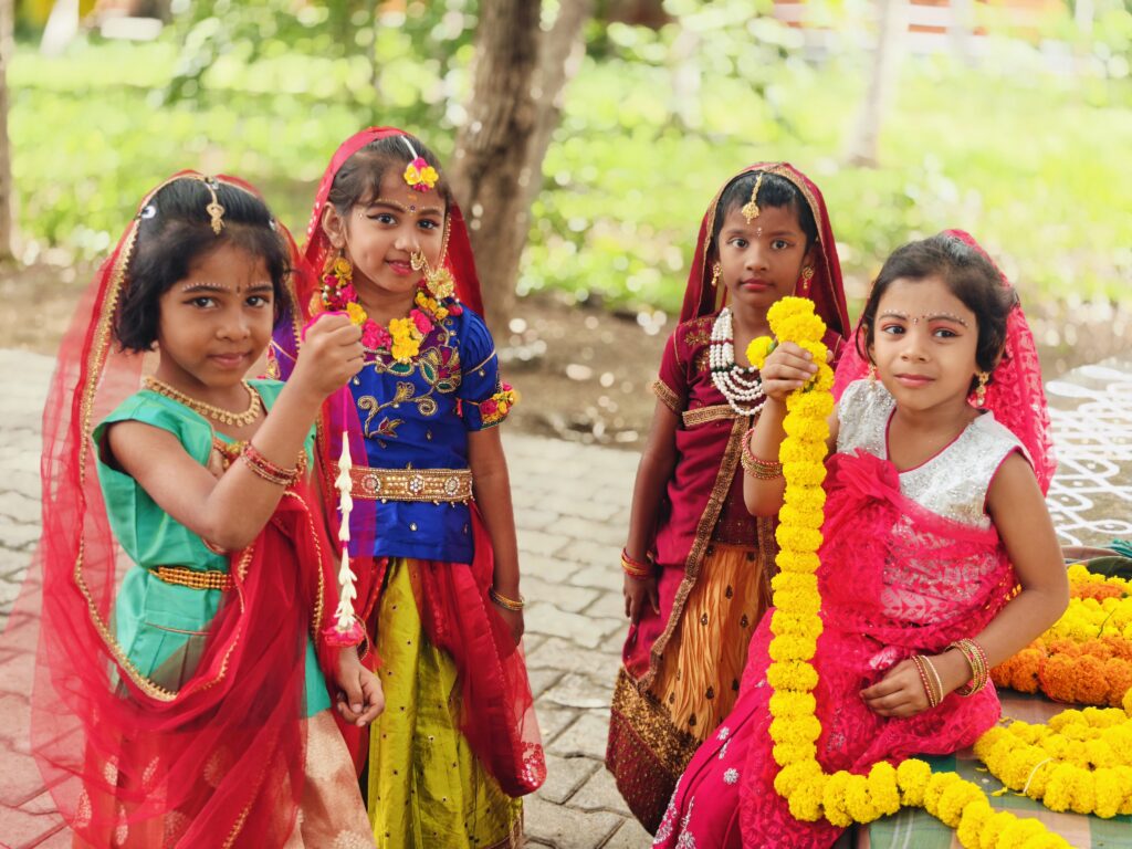 Four children in red veils and festive outfits hold marigold garlands beside a pile of flowers.